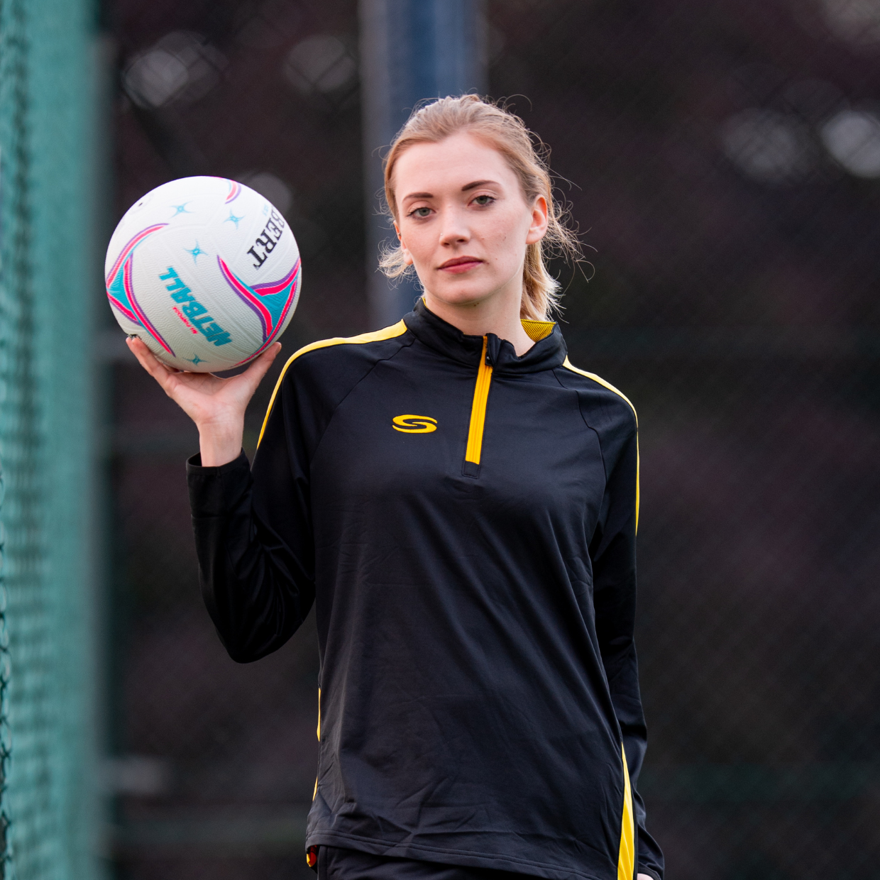 Teaser for Our Netball Teamwear Solution Netball player in black/yellow midlayer holding a netball