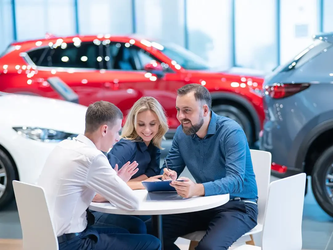 A couple sitting at a table speaking with a car dealer with new cars behind them