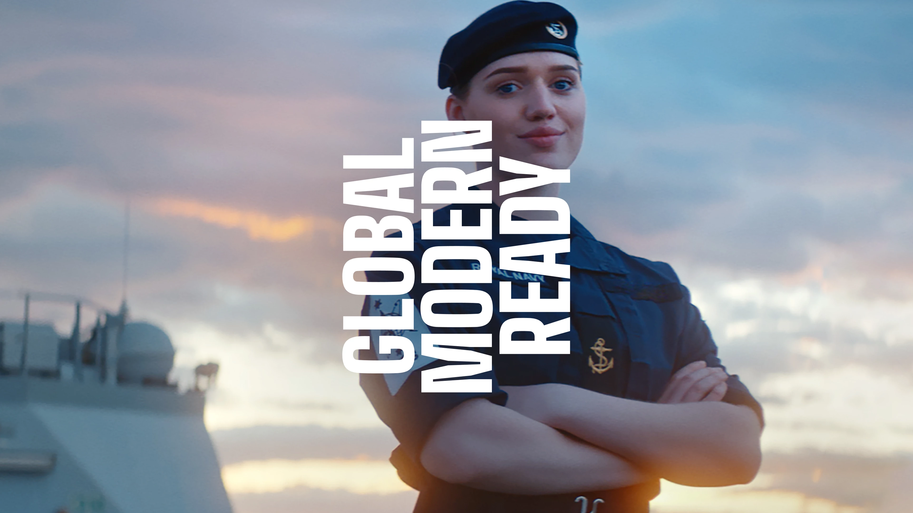 A female member of the Royal Navy stands in front of a ship with the words Global, Modern, Ready in front of her