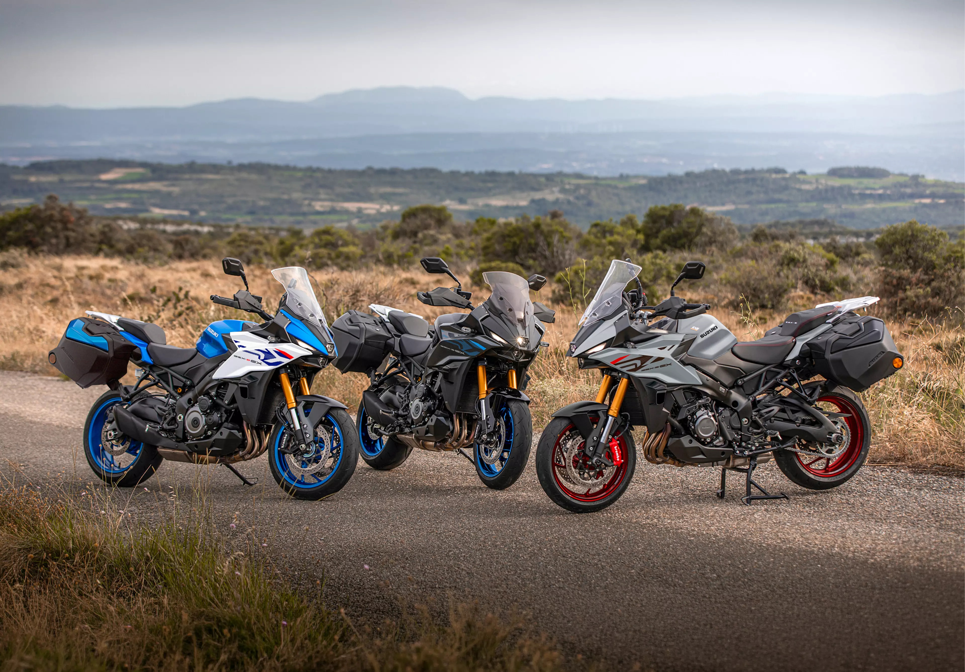 Suzuki GSX-S1000GX+ three colour line-up with bikes static and a wood in the background.