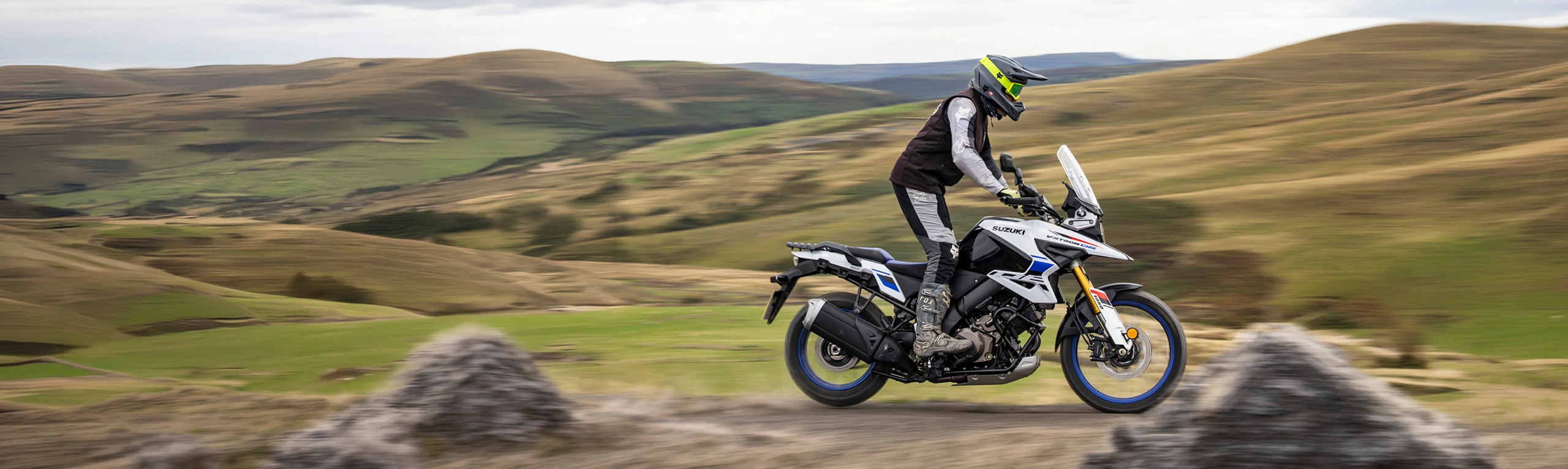 Suzuki V-Strom 1050DE white, side on pushing on along a trail on top of a valley
