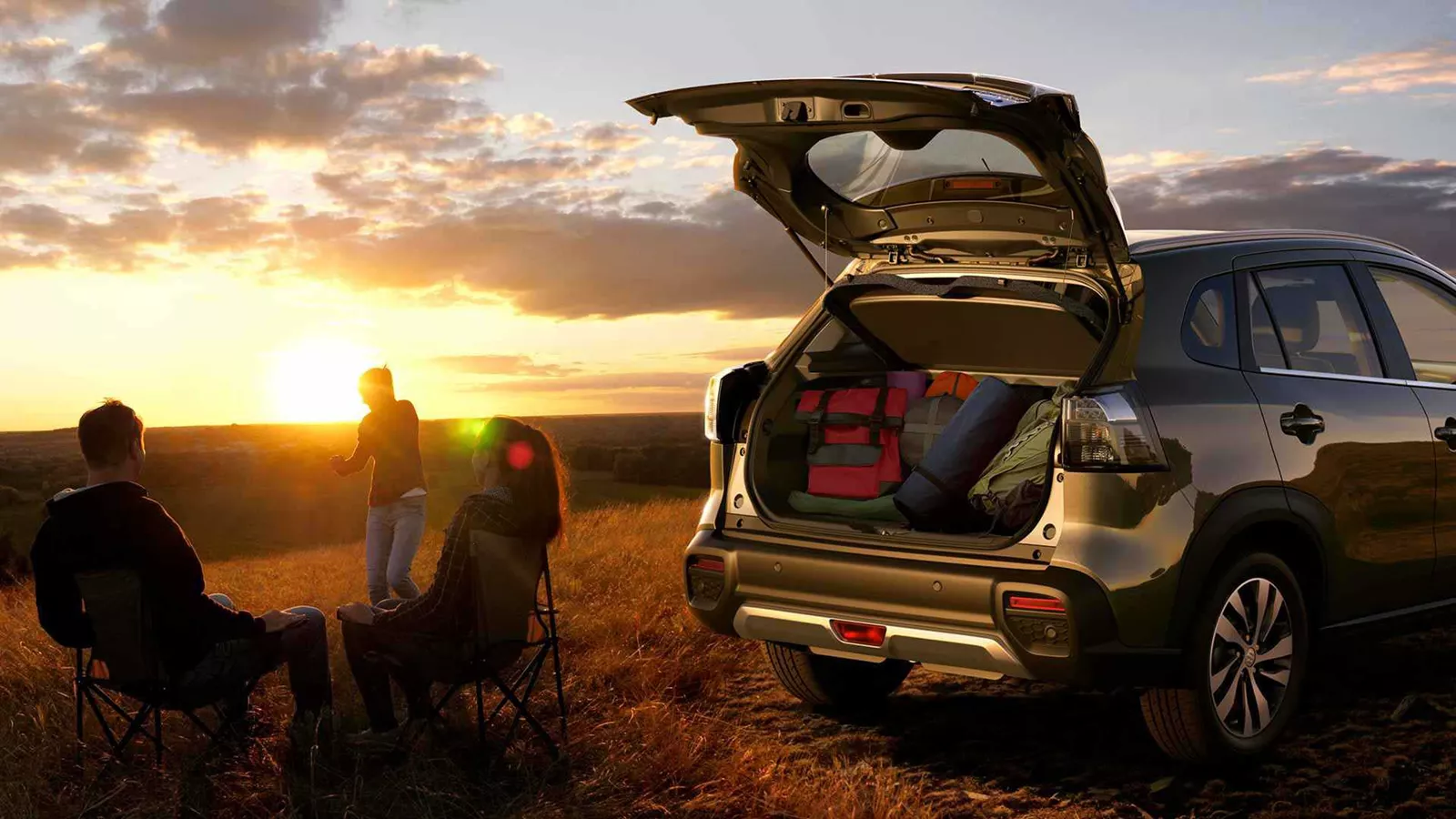 Suzuki e Vitara parked at sunset with open boot packed for a trip, as three people relax outdoors in a countryside setting