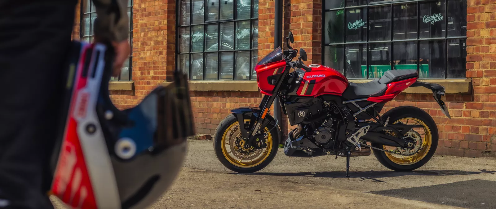 Suzuki GSX-8TT red with a rider holding the helmet to the left and the bike left hand side on in front of an old industrial building