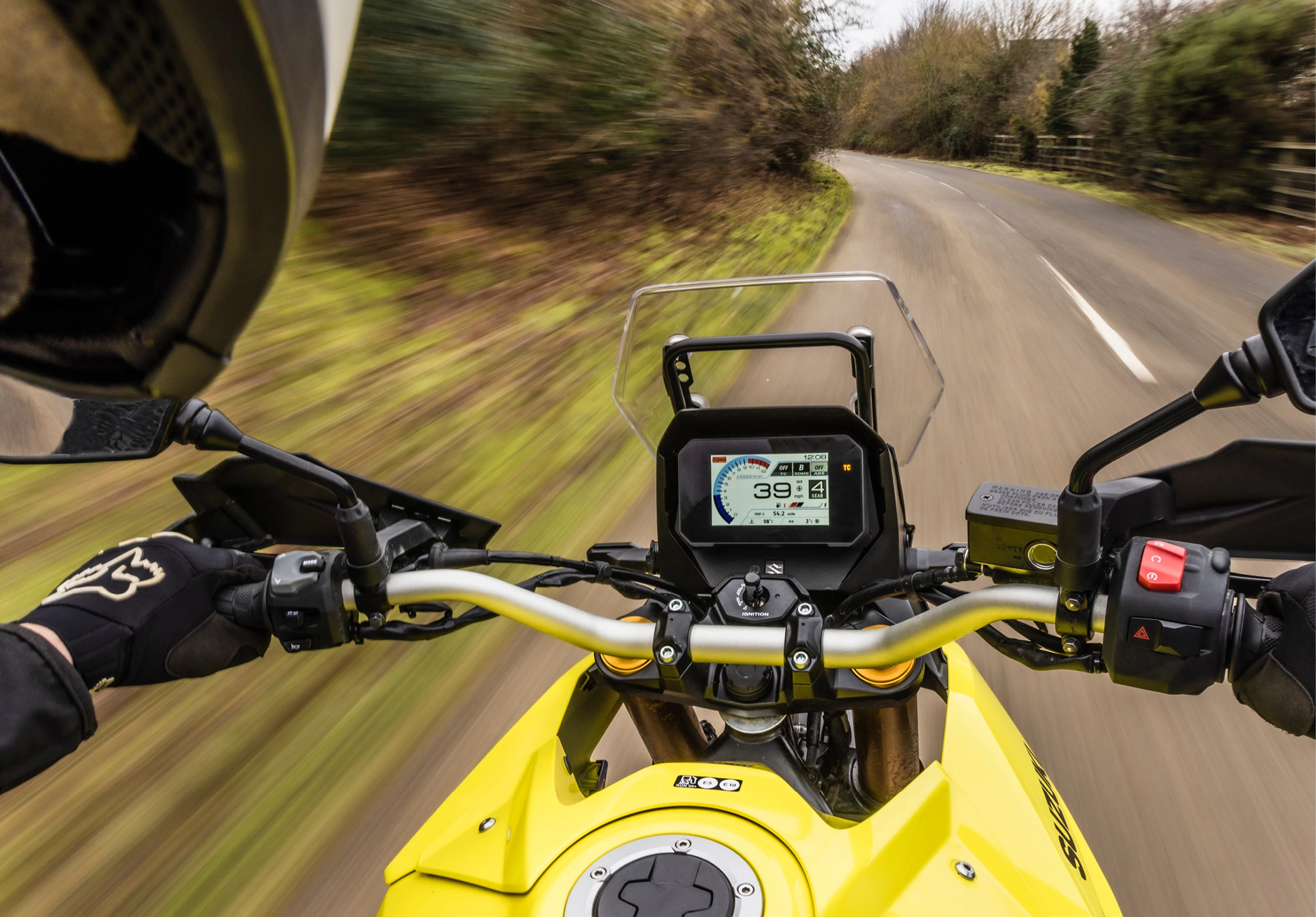 A rider’s perspective of the Suzuki V-Strom 800DE’s digital instrument cluster, showing the bike in motion on a winding road.