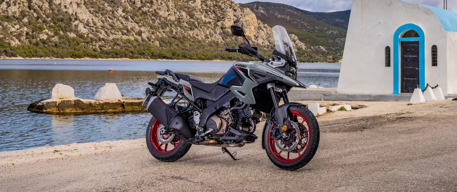 Suzuki V-Strom 1050RE grey, static beside a church with a river and mountain backdrop