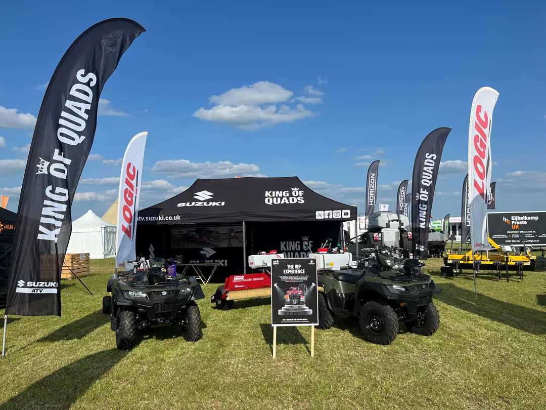 Outdoor Suzuki King of Quads display at an agricultural show, featuring branded flags, a black Suzuki marquee, quad bikes, trailers, and promotional signage on a grassy exhibition stand