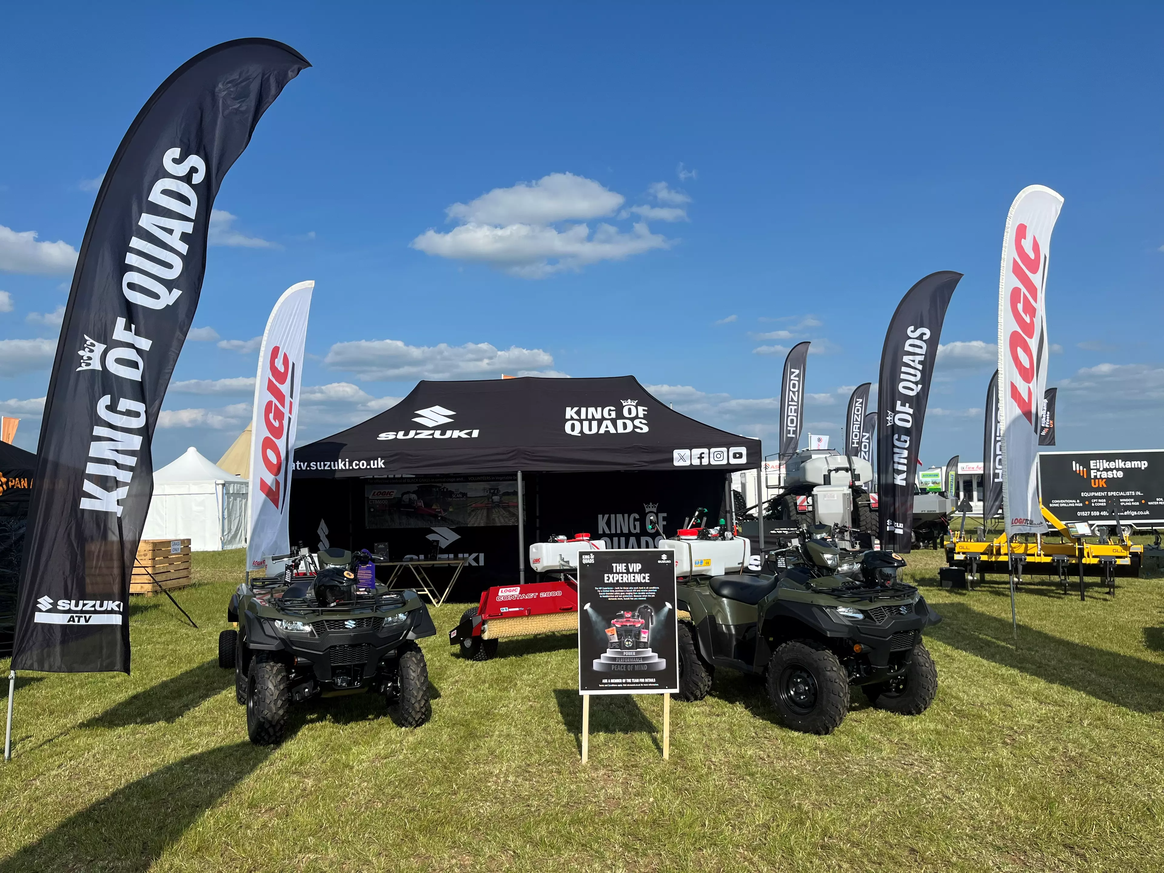 Outdoor Suzuki King of Quads display at an agricultural show, featuring branded flags, a black Suzuki marquee, quad bikes, trailers, and promotional signage on a grassy exhibition stand