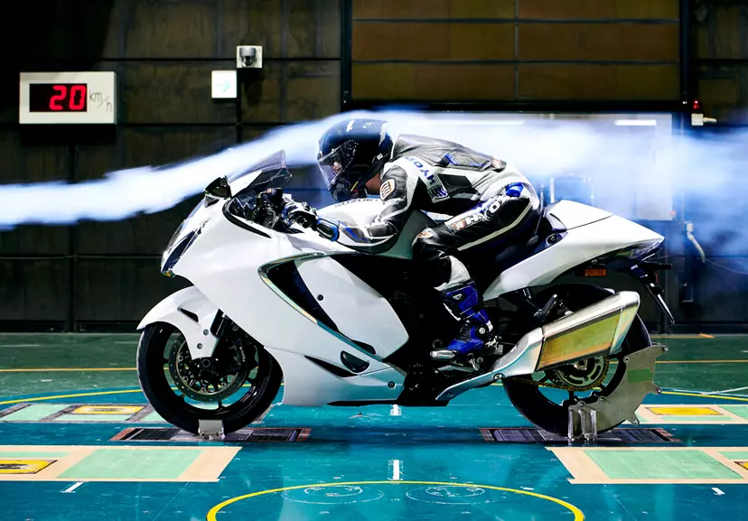 Suzuki Hayabusa motorcycle in a wind tunnel testing facility with rider in aerodynamic position and smoke trails showing airflow.