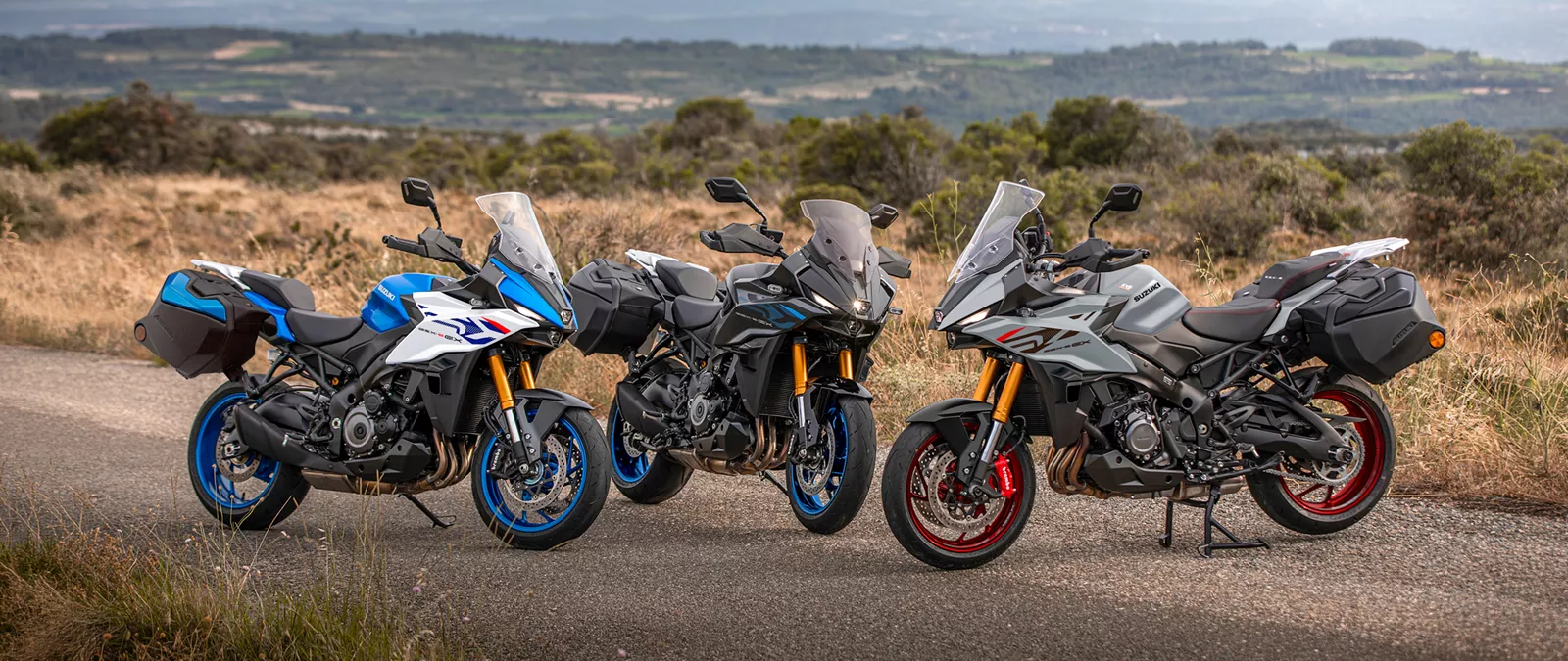 Suzuki GSX-S1000GX+ three colour line-up with bikes static and a wood in the background.