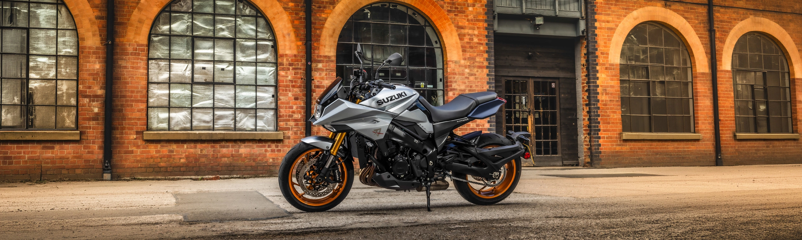 Suzuki Katana silver left hand side static with a brick building with large windows behind