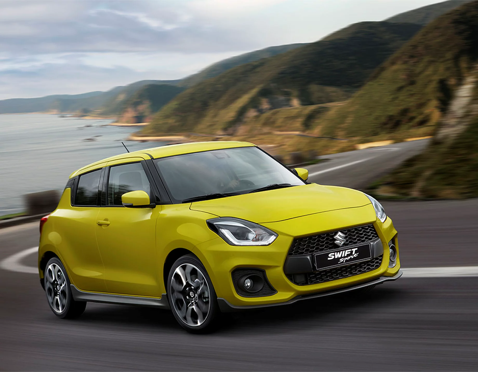 Yellow Suzuki Swift Sport driving along a coastal road with the sea and hills in the background.