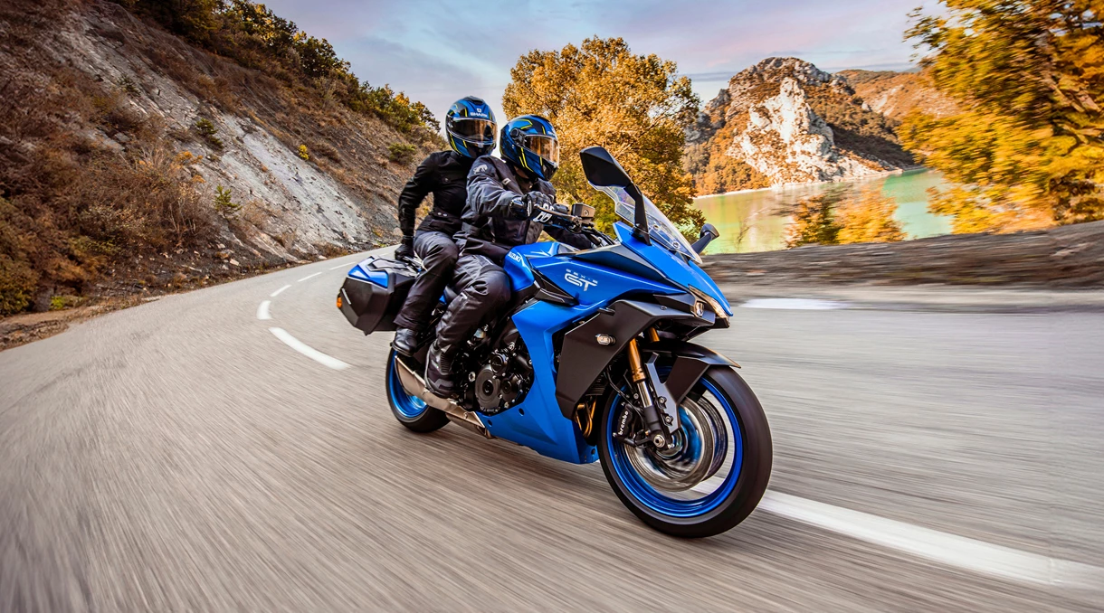 Suzuki GSX-S1000GT with a rider leaning slightly into a curve on a winding road, surrounded by a scenic mountain landscape