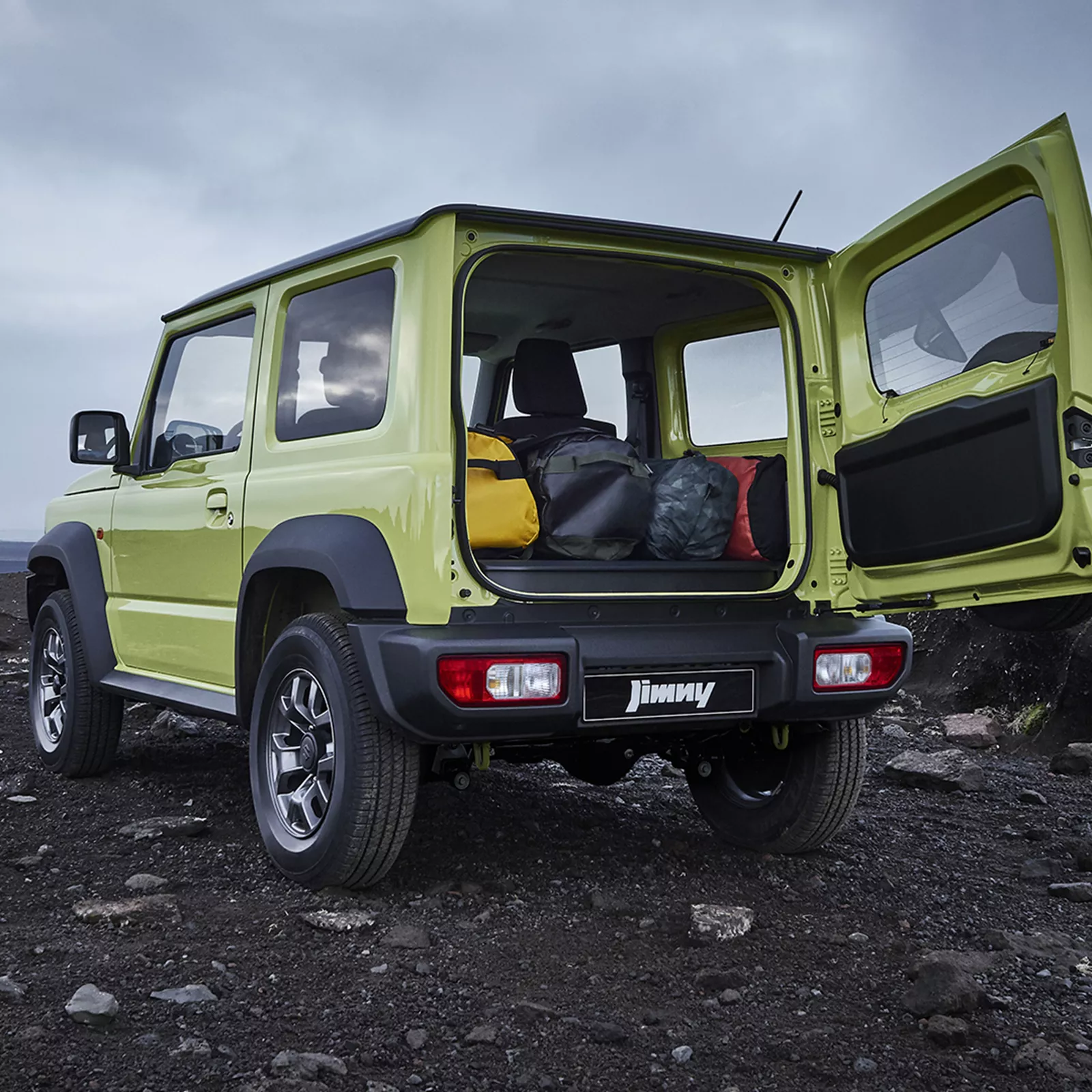 Rear view of Suzuki Jimny with boot open, showcasing luggage space and rugged off-road setting.