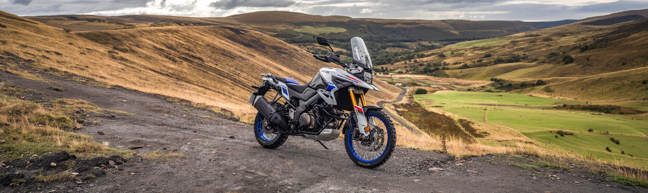 Suzuki V-Strom 1050DE white, side on front three quarters on top of a hill with a valley behinf