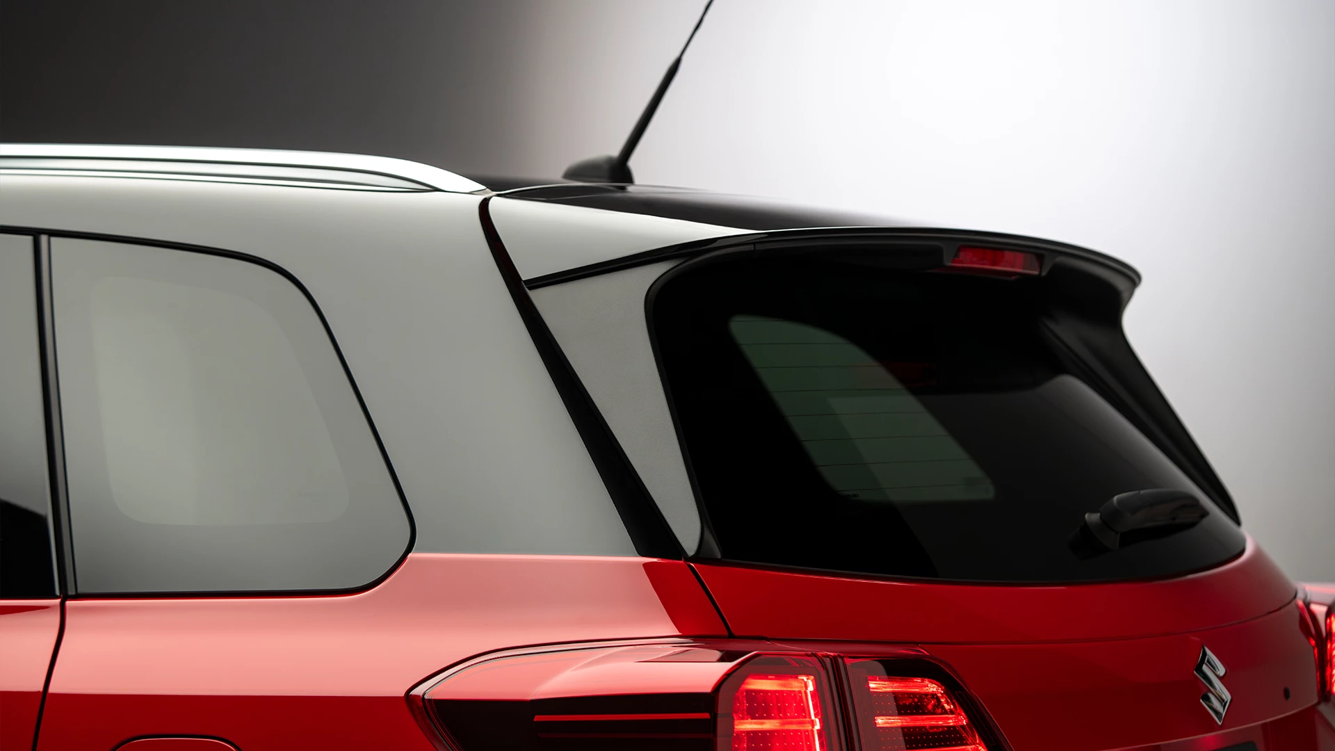 Close-up of the rear upper section of a bright red Suzuki Vitara, highlighting the roof spoiler, cosmic black roof, privacy glass and rear antenna.