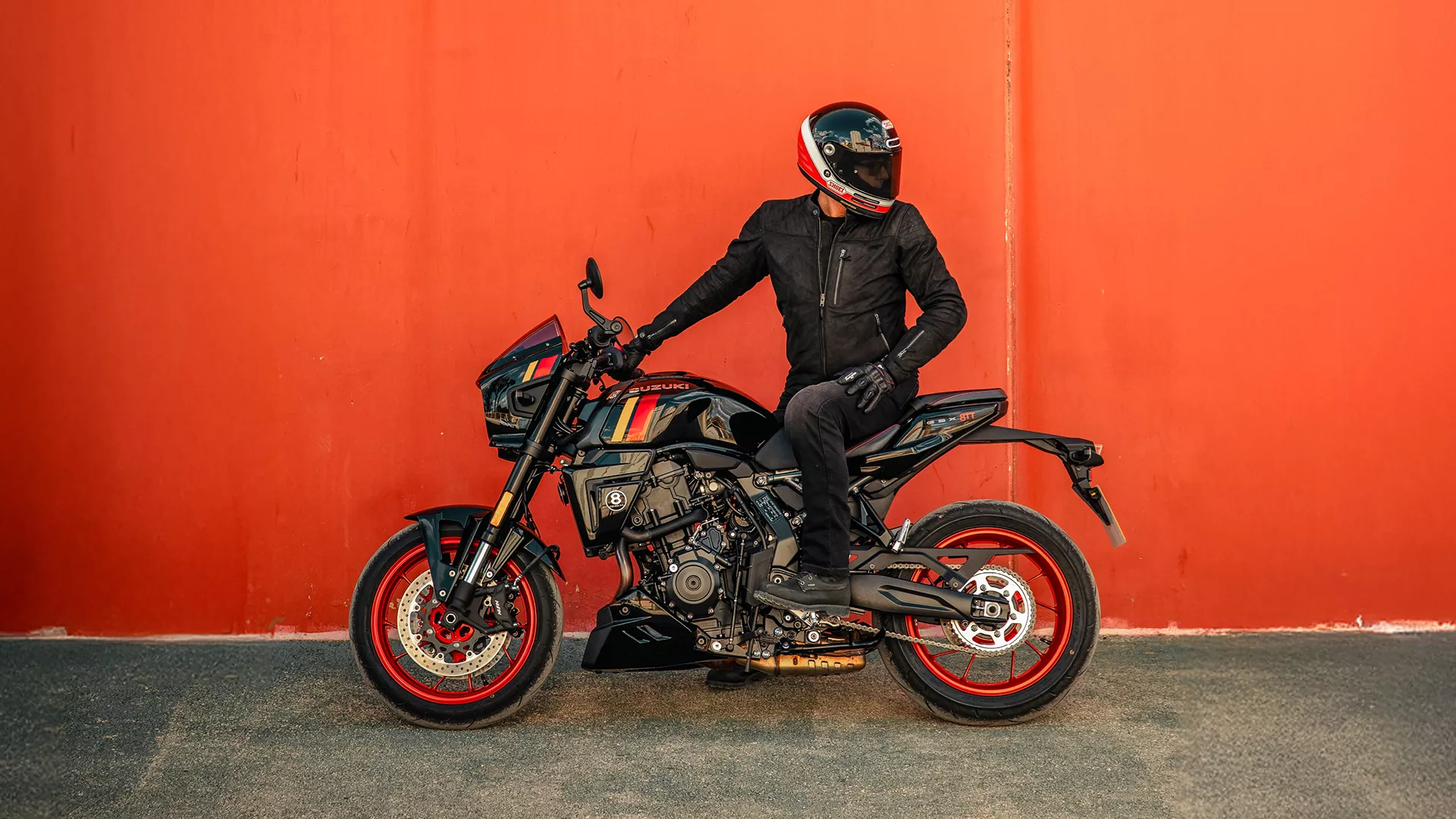Suzuki GSX-8TT black, static with the rider looking back at their bike with a red wall behind