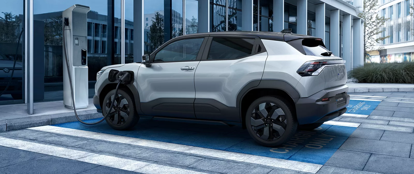 Suzuki e-Vitara plugged into a city centre EV charging station beside a modern glass office building