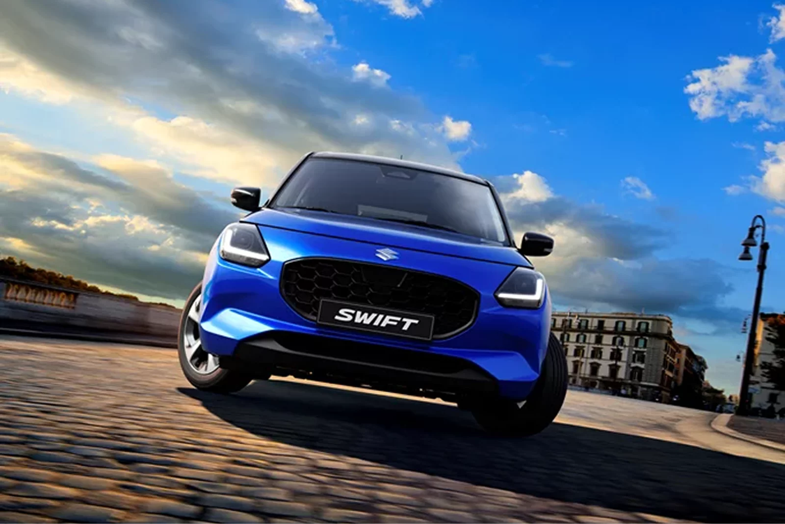 Front-facing view of a blue Suzuki Swift driving on a cobbled city street