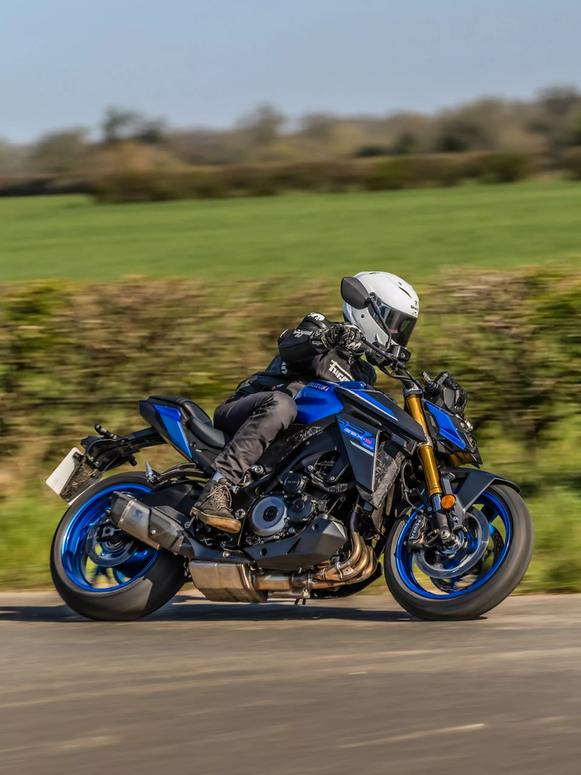 Suzuki GSX-S1000 leant over into a corner with fields and a hedge behind.