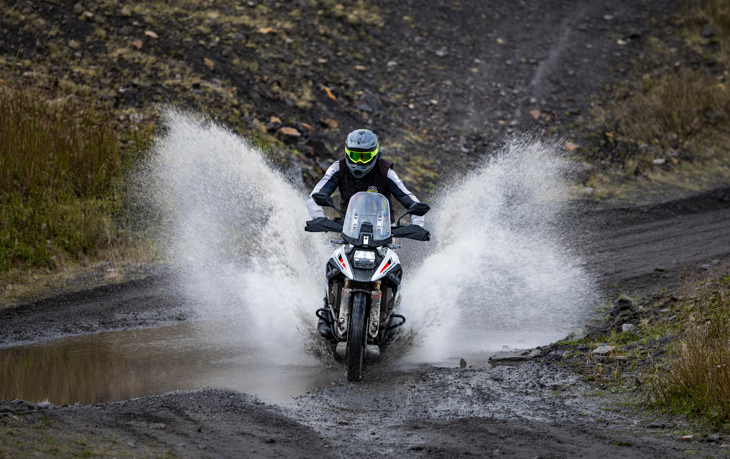 Suzuki V-Strom 1050DE white moving through a water splash on a mountain trail