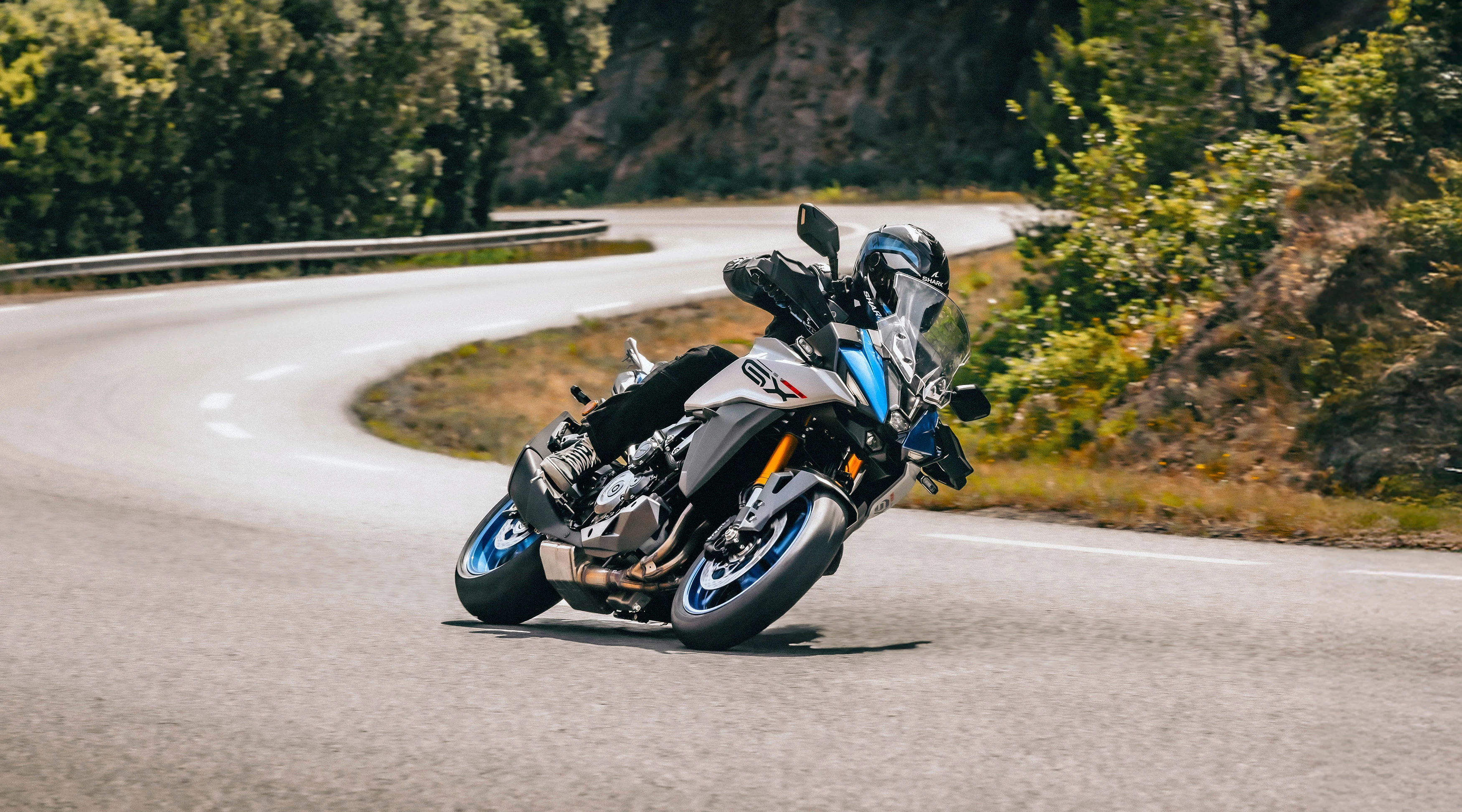 Suzuki GSX-S1000GX motorcycle taking a sharp turn on a road, showcasing its agility and performance.