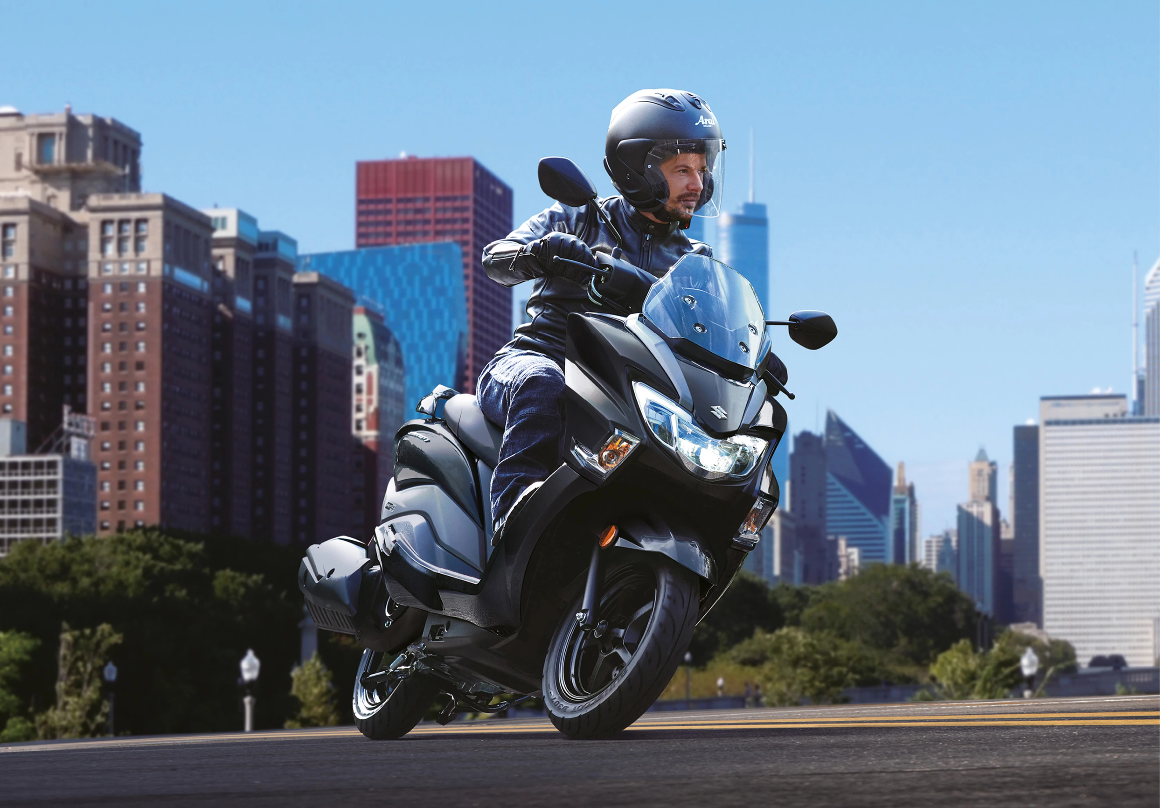 ASuzuki Burgman Street 125EX rider in a black leather jacket riding past a modern glass building, highlighting the scooter’s stylish urban appeal.