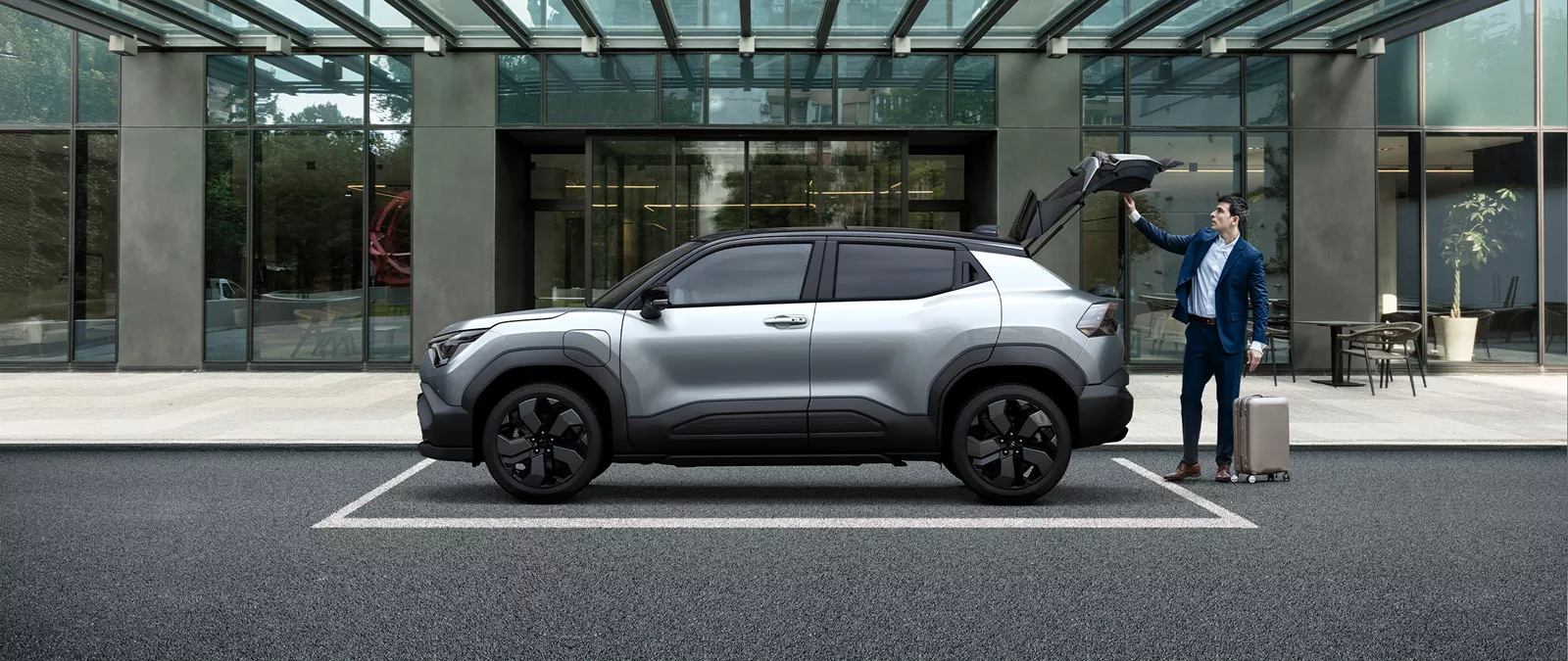 Man loading luggage into the boot of a Suzuki e-Vitara parked outside a modern glass-fronted building