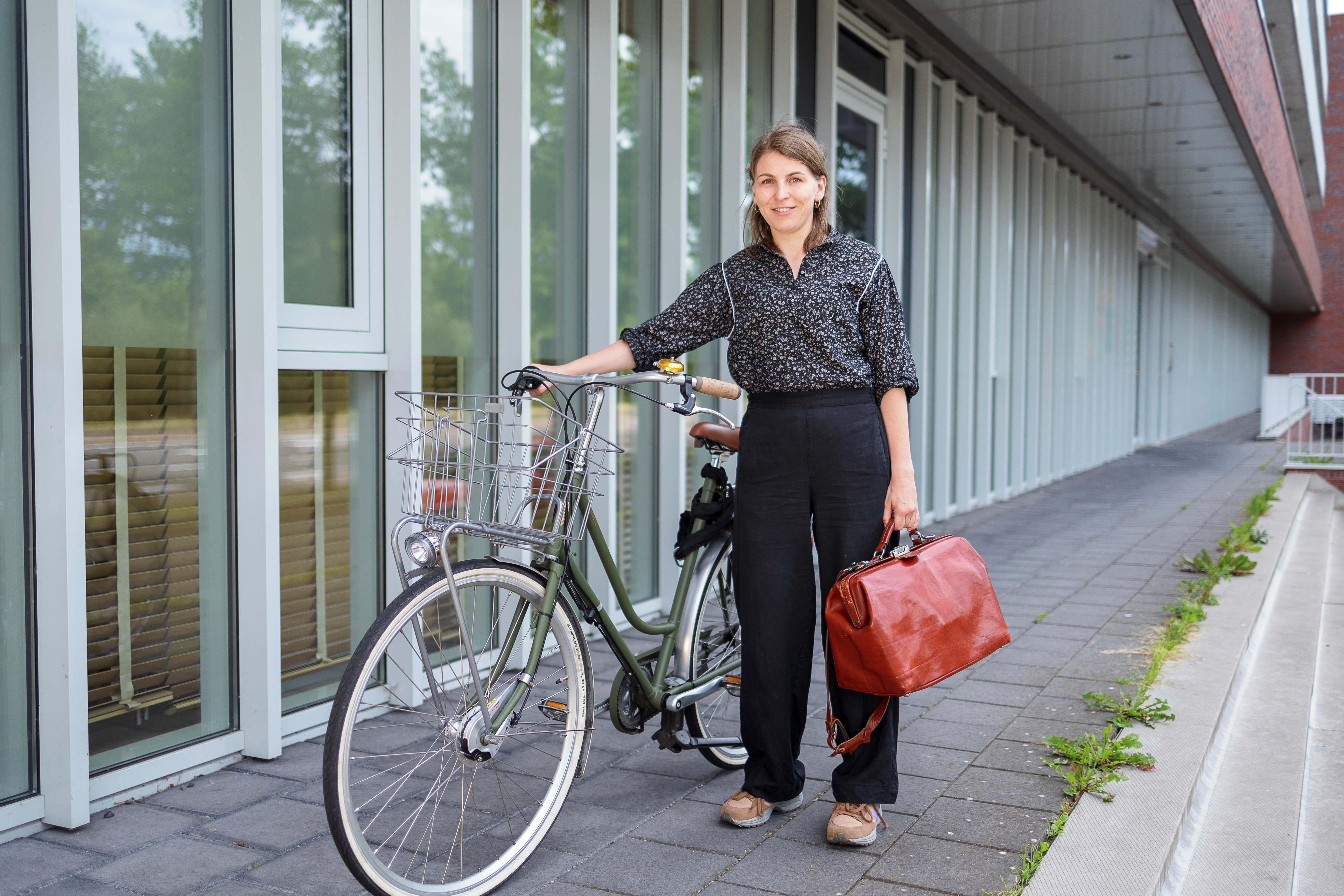 Vrouw met dokterstas in hand houdt fiets vast en kijkt glimlachend in de camera