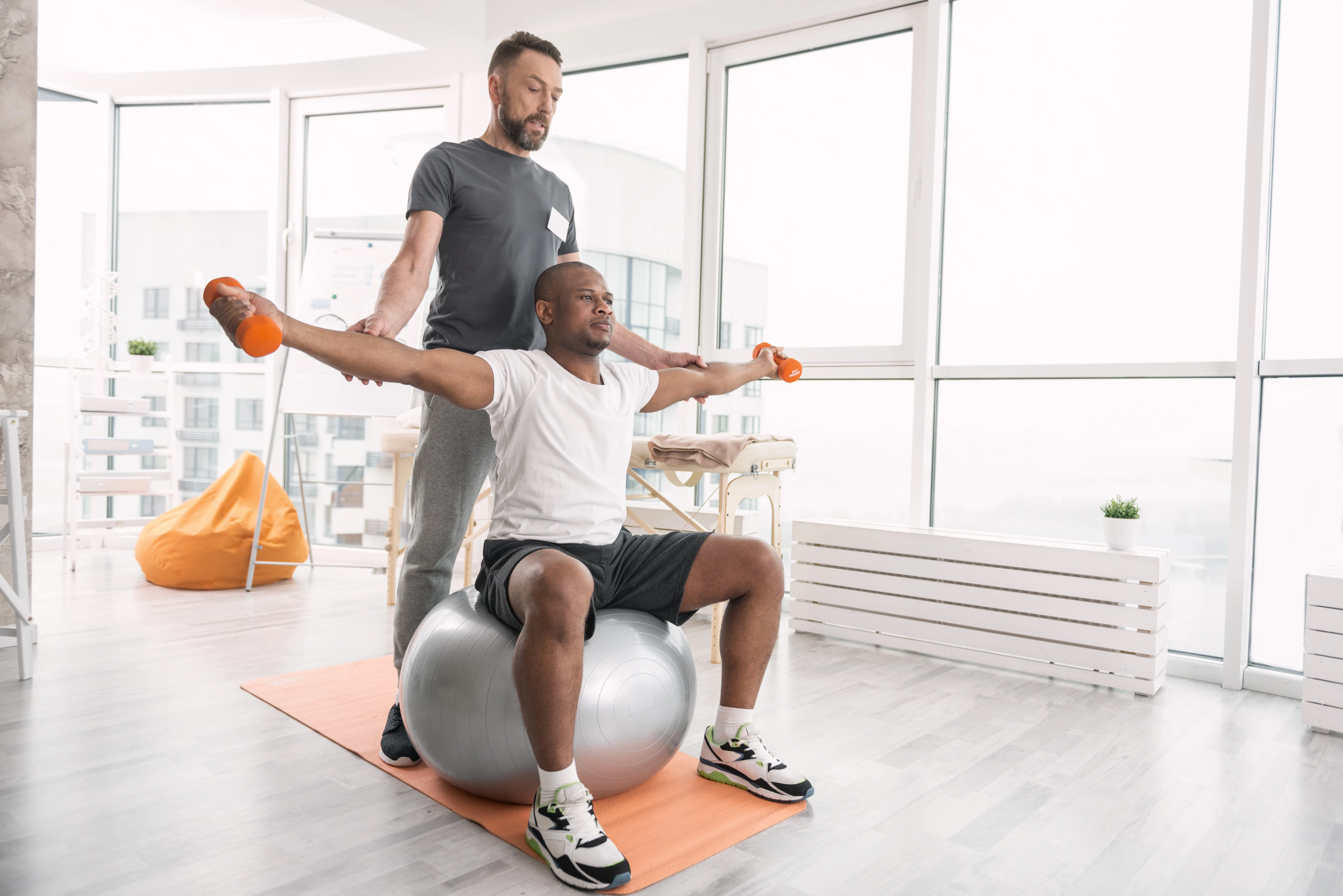 Een man zit op een skippybal en heeft met gestrekte armen gewichtjes vast en een andere man helpt hem.