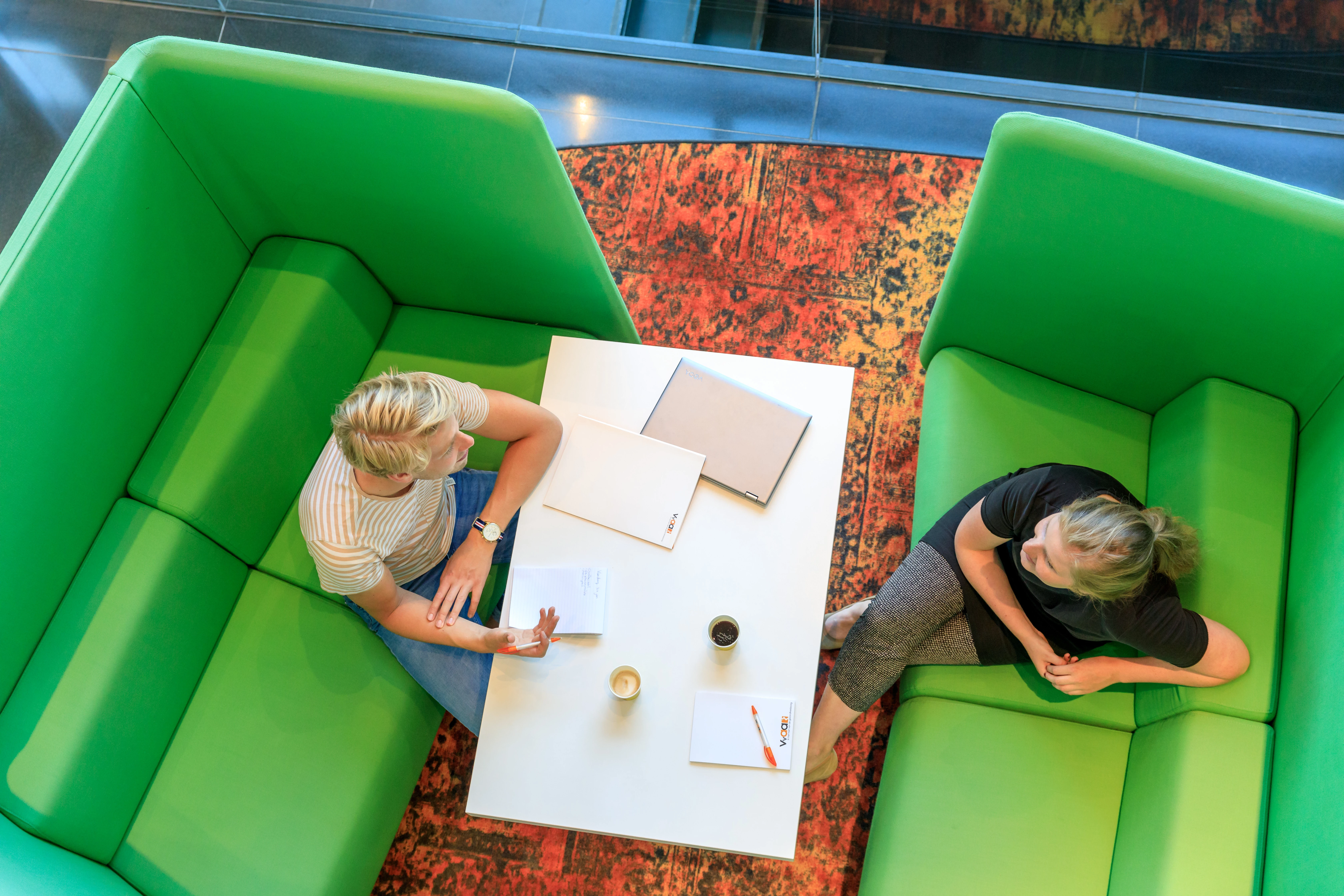 Een man krijgt coaching aan een witte tafel op groene banken van een vrouw die achterover geleund zit.