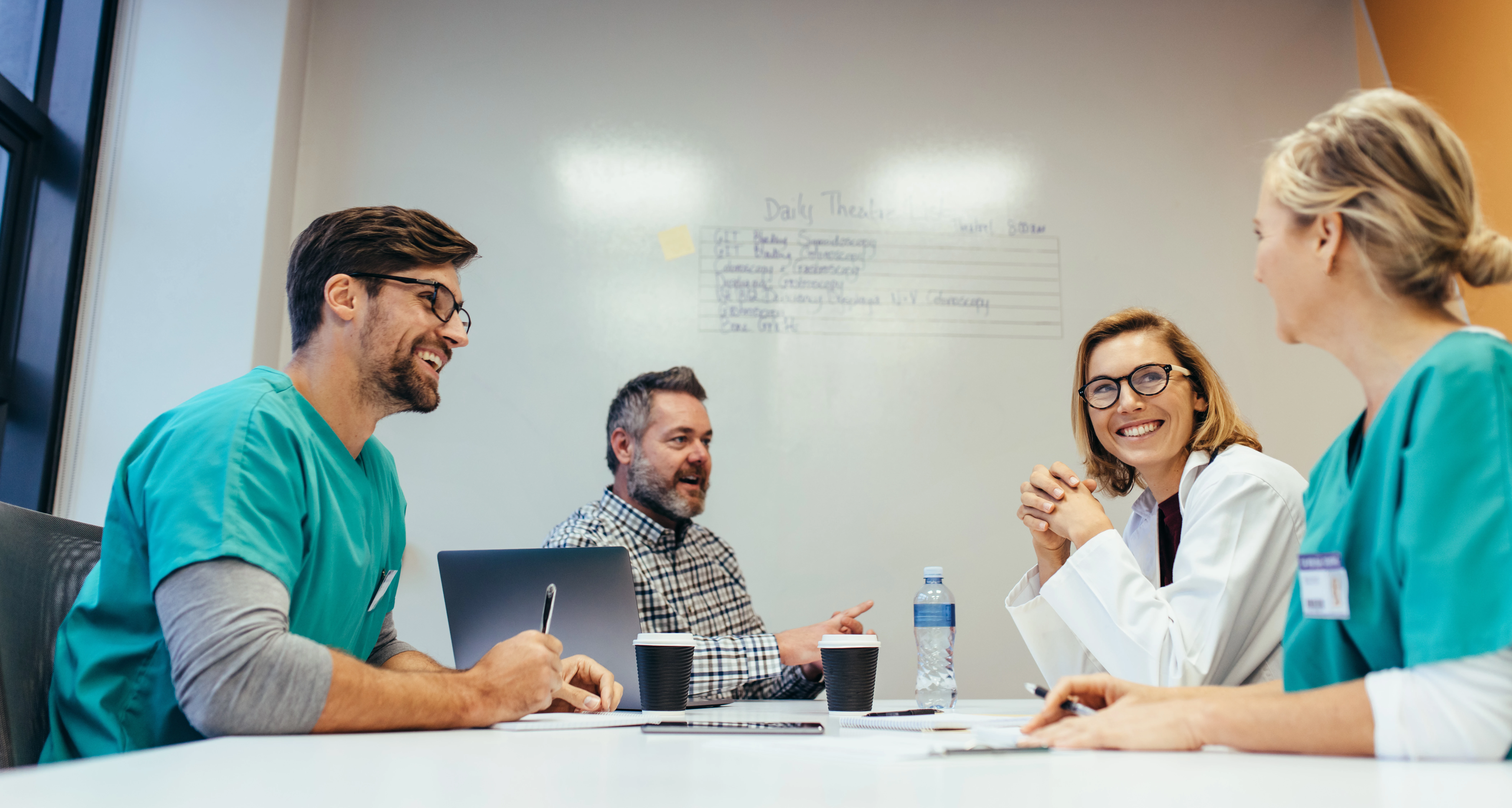 Vier mensen zitten aan een tafel met kopjes koffie, een flesje water, een laptop en pen en papier.