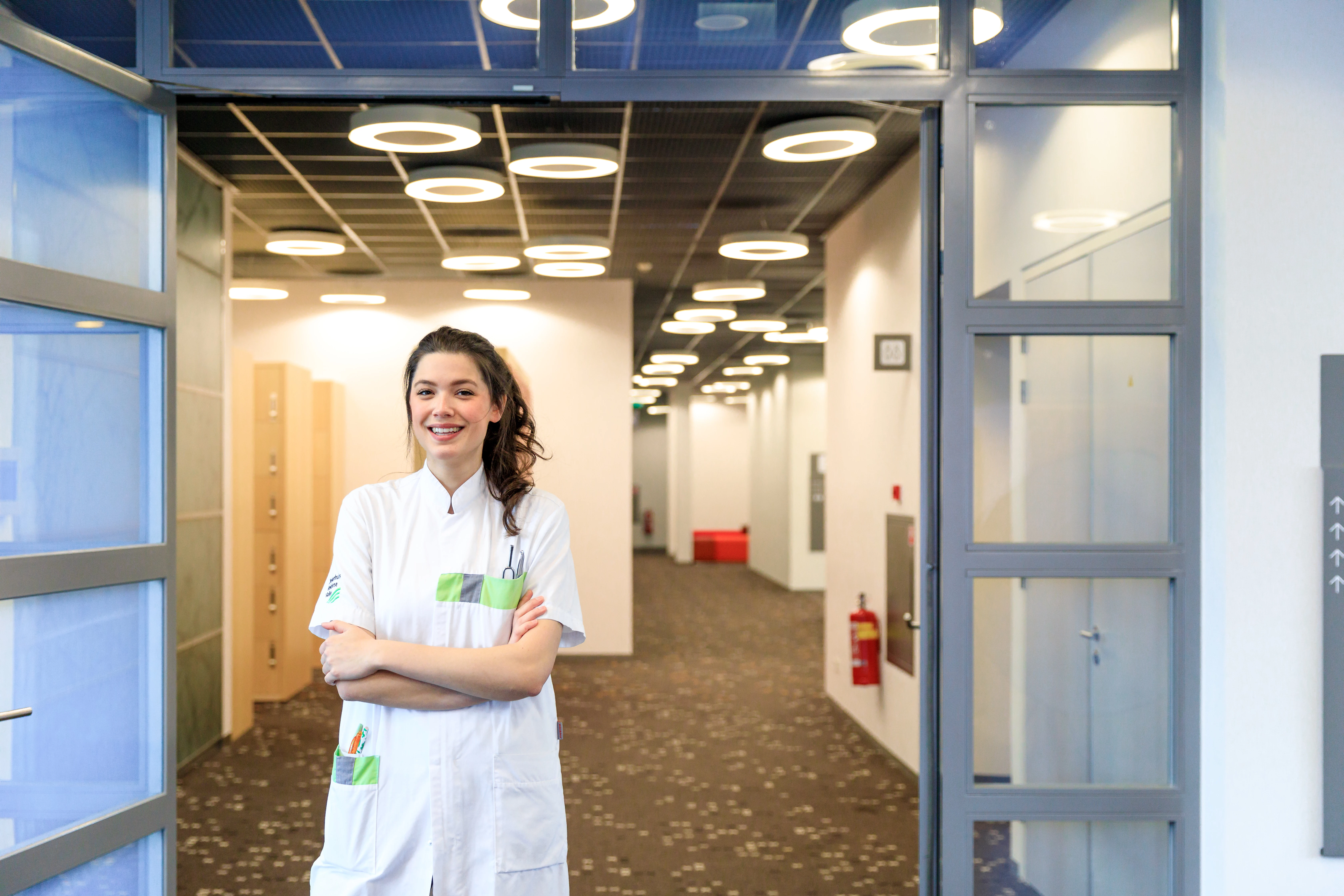 Meisje met zorgverlenersoutfit lacht met armen over elkaar en staat in de gang
