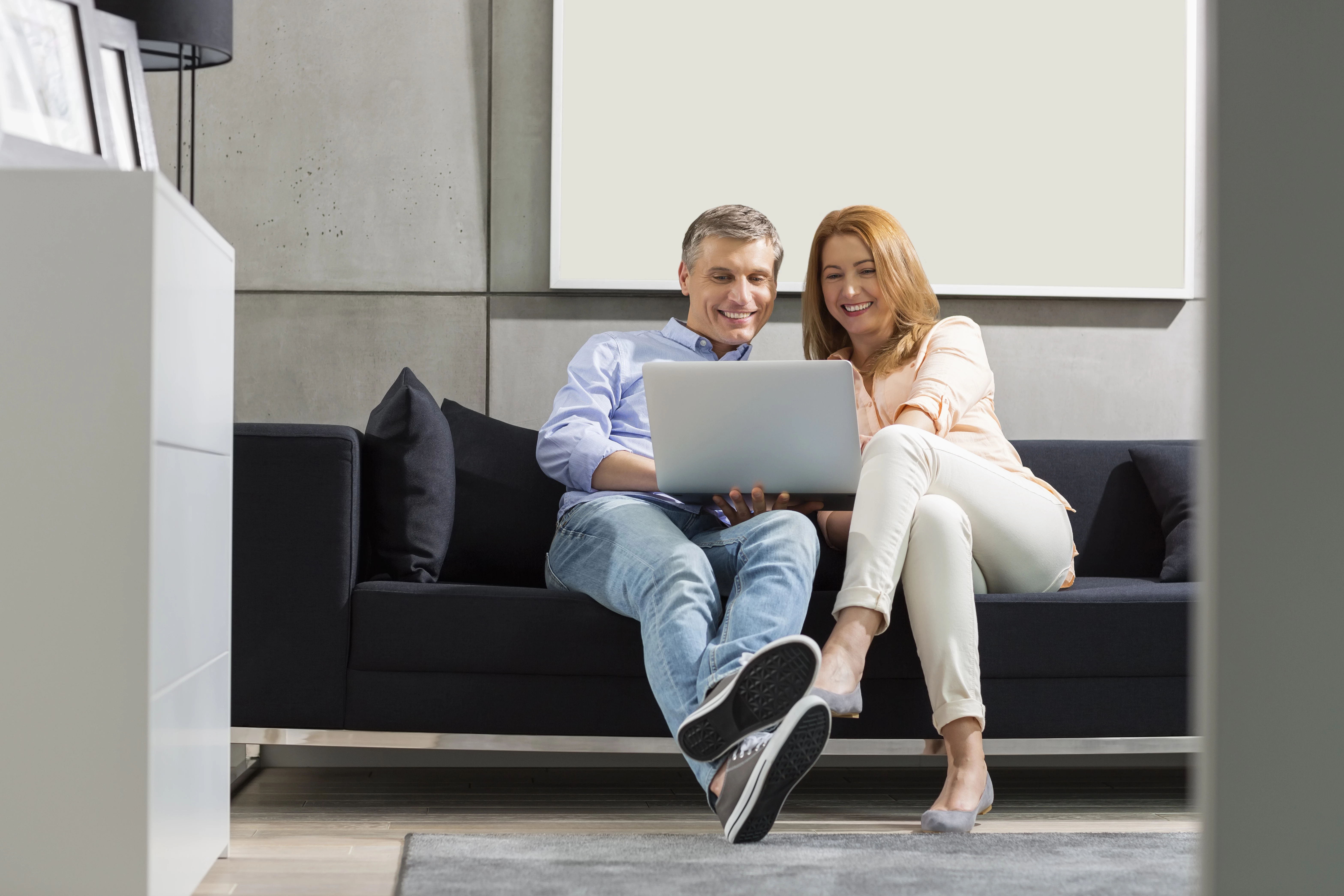 Een man en een vrouw zitten op een zwarte bank en lachen naar de laptop op hun schoot.