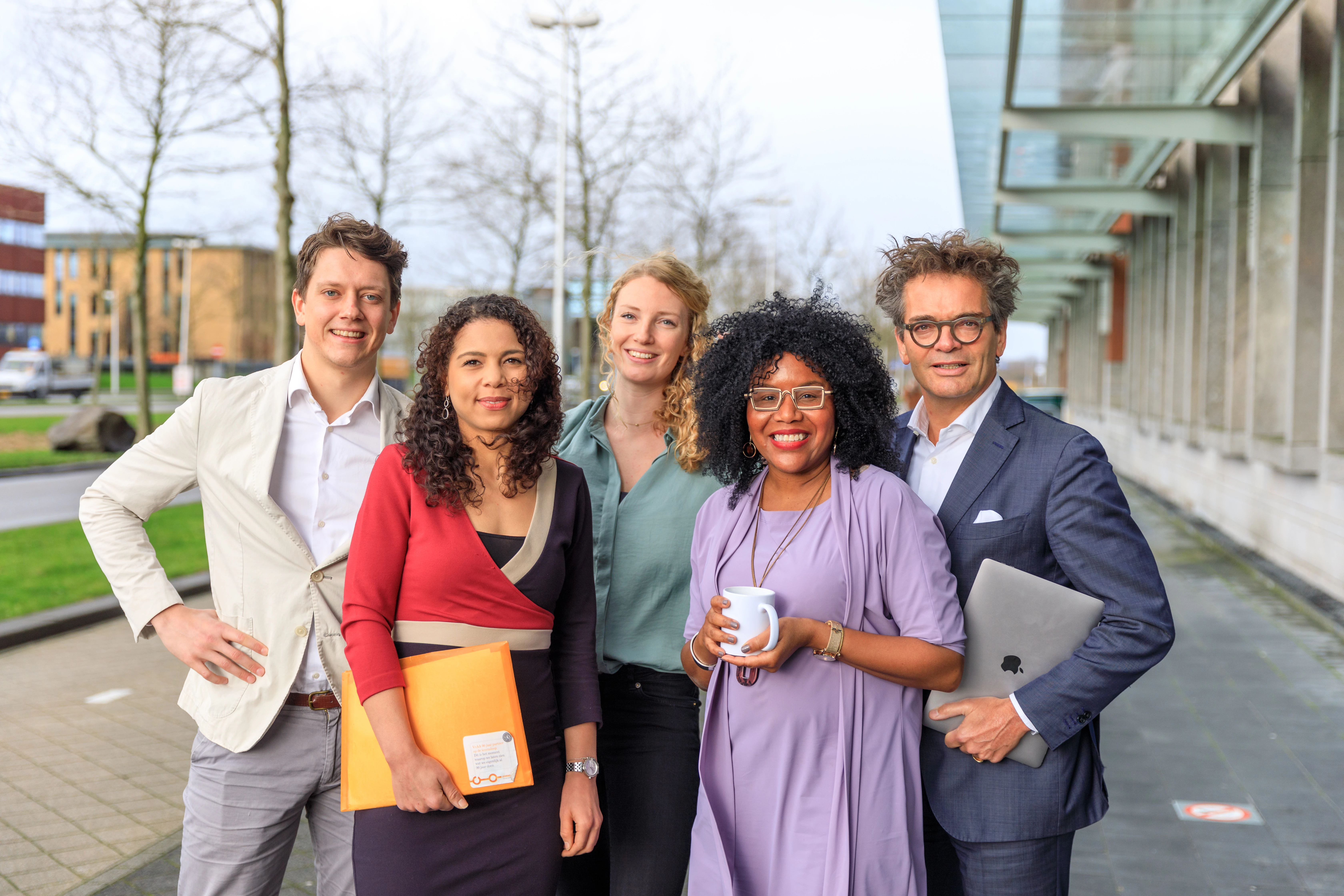 Vijf personen staan met een laptop, mapje en kopje buiten en lachen naar de camera.