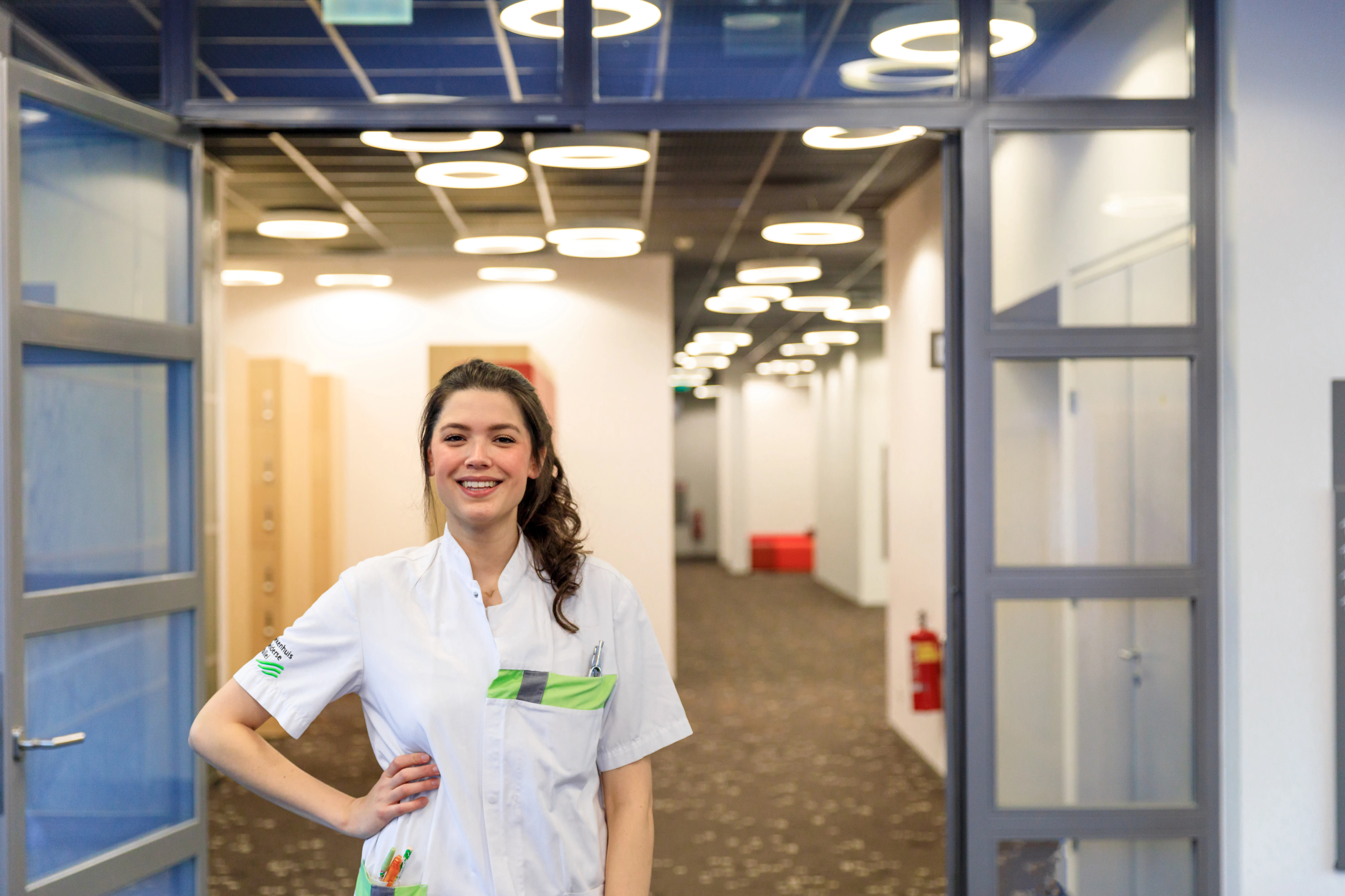 Een vrouw met een wit pak staat met een arm in haar zij in een gang