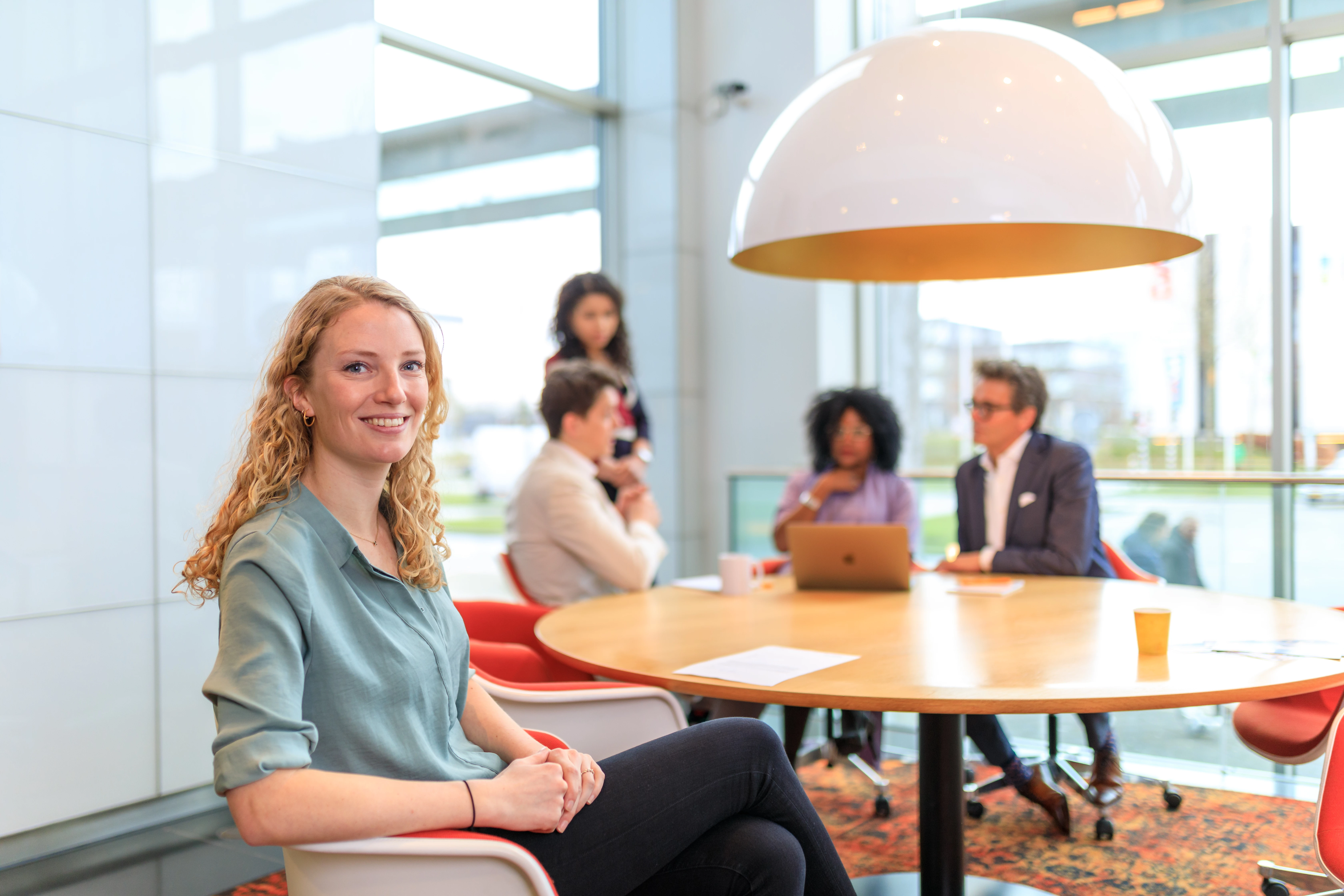 Een vrouw met blonde krullen zit op een stoel en op de achtergrond is een teamcoach met drie personen aan het praten.