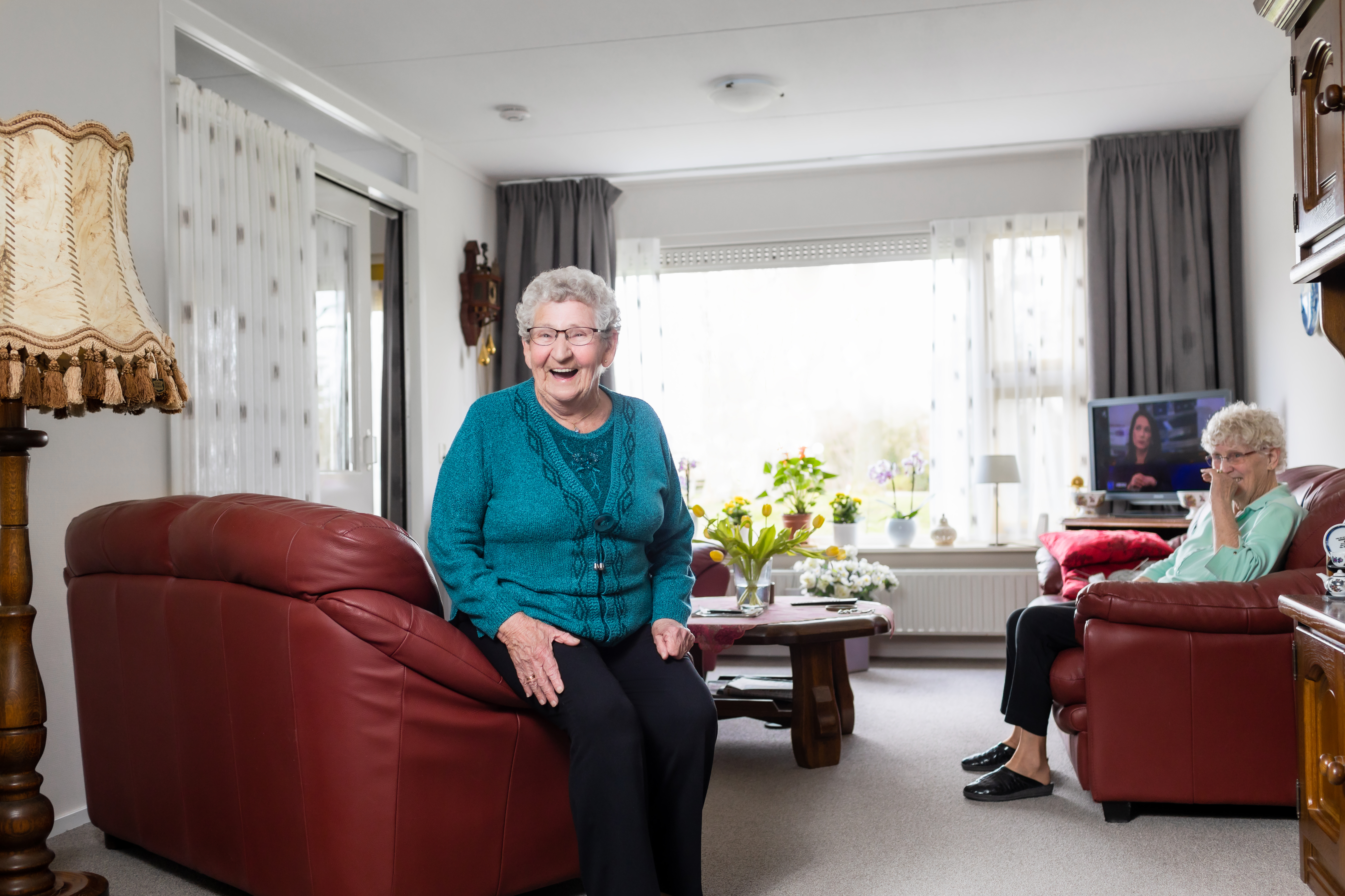 Oudere vrouw kijkt vanuit haar huiskamer lachend op een leren stoel in camera
